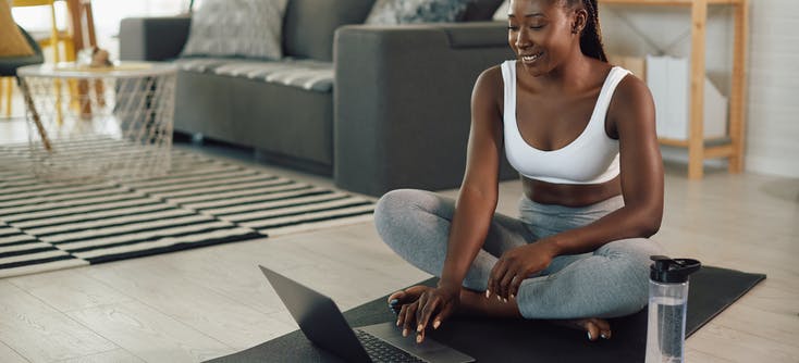 A fitness instructor sitting on an exercise mat and working on her laptop.